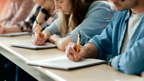 Filmfootage-aus-einer-Reihe-von-Multi-ethnischen-Schüler-im-Klassenzimmer-Prüfung-/-Test-Holding-Stifte-und-Schreiben-in-Notebooks-im-Fokus.-Helle-Jugendliche-Studie-an-der-Universität.