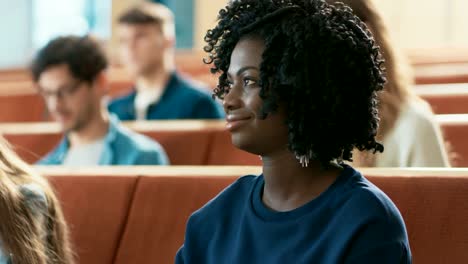 Close-up-of-a-Beautiful-Black-Female-Student-Sitting-Among-Her-Fellow-Students-in-the-Classroom,-She's-Writing-in-the-Notebook-and-Listens-to-a-Lecture.