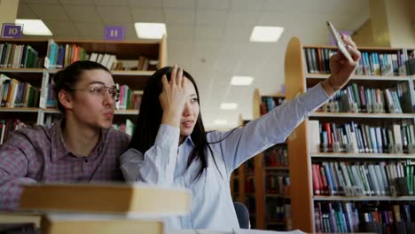 Junge-Teenager-paar-Studenten-sitzen-am-Tisch-in-der-Bibliothek-umgeben-von-Büchern,-die-immer-bereit-für-die-Prüfung.-Mädchen-hält-ihre-Smartpone-und-nehmen-Selfie-mit-ihrer-Klassenkameradin