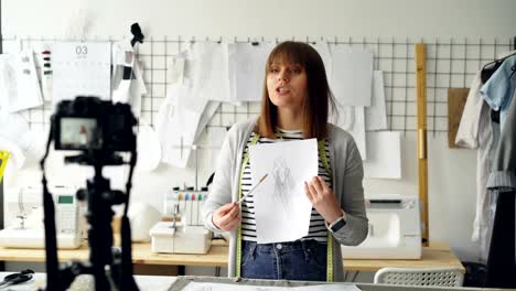 Young-blogger-seamstress-is-recording-video-for-instagram-about-designing-women's-clothing.-She-is-greeting-followers,-showing-sketch,-speaking-emotionally-and-gesturing.