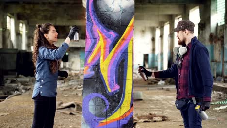 Two-skilled-graffiti-artists-bearded-guy-and-attractive-young-woman-are-working-together-in-abandoned-warehouse-decorating-old-column-with-abstract-image.