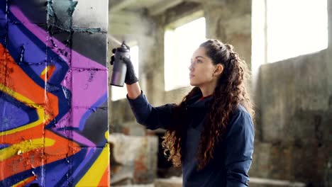 Serious-young-woman-graffiti-artist-is-painting-on-column-in-old-warehouse-using-spray-paint.-Abandoned-dirty-building-with-litter-on-floor-is-in-background.