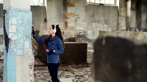 Tilt-Up-Schuss-des-modernen-urbanen-Malers-Zeichnung-Graffiti-auf-hohe-Säule-im-verlassenen-Lagerhaus-mit-Aerosol-Farbe-und-Musikhören-mit-Kopfhörer.