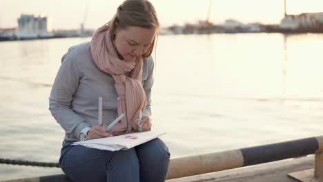 Junge-Frau-sitzt-in-der-Nähe-von-Pier-im-Frühjahr-Abend-und-Zeichnung-von-Bleistiften