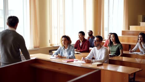 Experienced-high-school-teacher-is-talking-to-cheerful-students-while-young-people-are-listening,-smiling-and-laughing-sitting-at-desks-in-light-classroom.