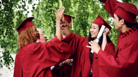 Multiracial-group-of-young-people-students-are-doing-high-five-on-graduation-day-wearing-traditional-clothes-hats-and-gowns.-Millennials,-youth-and-education-concept.