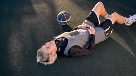 Beautiful-blonde-female-basketball-player-lying-on-court-and-listening-music,-woman-relaxing-in-open-air-during-sunset