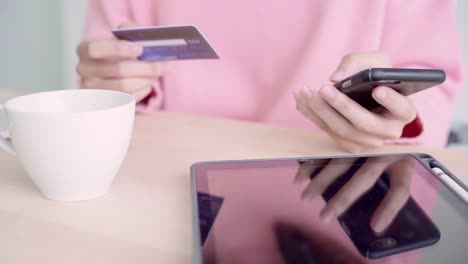 Beautiful-Asian-woman-using-smartphone-buying-online-shopping-by-credit-card-while-wear-sweater-sitting-on-desk-in-living-room-at-home.-Lifestyle-woman-at-home-concept.