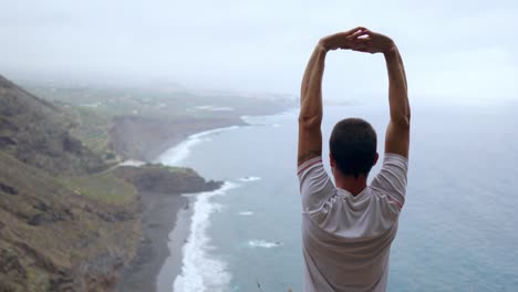 Ein-Mann-steht-am-Rand-einer-Klippe-mit-Blick-auf-den-Ozean-erhebt-seine-Hände-und-atmet-die-Luft-während-yoga