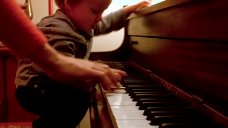 Caucasian-Toddler-Boy,-Sitting-at-a-Piano-with-Him-Mom,-Plays-on-the-Piano-with-a-decorated-Christmas-Tree-Behind-Them