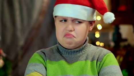 The-boy-in-the-sweater-and-the-hat-of-Santa-Claus-is-upset-and-offended.-He-has-no-gift.-Close-up,-against-the-background-of-Christmas-lights