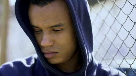 Upset-afro-american-guy-standing-near-metal-grid-of-military-camp,-feeling-sad