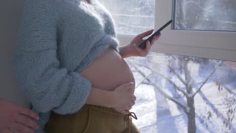 moderne-Elternschaft,-Mann-Hände-streicheln-Bauch-der-schwangeren-Frau-mit-Handy-in-Nahaufnahme-Arm-in-natürliches-Licht-in-der-Nähe-von-Fenster