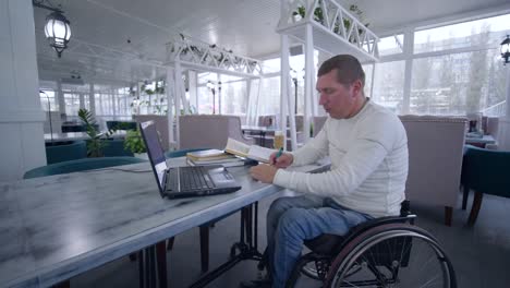 online-education,-student-disabled-man-on-wheelchair-uses-modern-laptop-technology-to-learn-from-online-lessons-and-books-making-notes-in-notebook-close-up-sitting-at-table-in-cafe