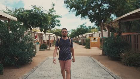 Bearded-tourist-man-is-walking-on-yard-of-resort-area-with-small-summer-cottages