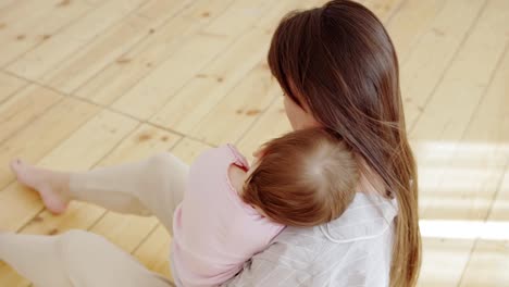 Vista-de-ángulo-alto-de-hermosa-madre-joven-sentada-en-el-piso-de-madera-en-casa-y-adormando-a-su-niña-linda-para-dormir