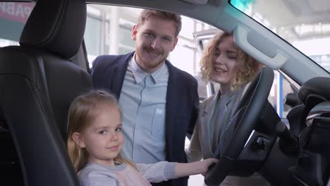 Lächeln-kleine-Mädchen-hinter-dem-Steuer-des-neuen-Fahrzeugs-zusammen-mit-Mama-und-Papa-beim-Kauf-von-Familienauto-beim-Händler