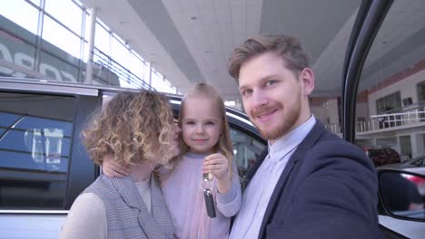 Konsumentfamilie-mit-niedlichen-kleinen-Tochter-mit-Schlüsseln-fotografieren-Telefon-in-der-Nähe-von-neu-gekaufter-Maschine-im-Auto-Showroom