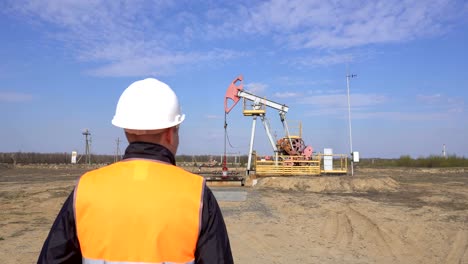 Un-joven-ingeniero-de-trabajo-en-un-chaleco-de-señal-endereza-un-casco-protector-en-su-cabeza-contra-el-fondo-de-una-máquina-de-mecedor-de-petróleo-y-gas,-una-industria,-espacio-de-copia,-pumpjack