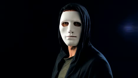 Anonymous-man-in-a-white-mask-covering-his-face-and-a-black-hood-turns-and-looks-at-the-camera.-Black-dark-background.-Concept-of-a-member-of-a-secret-organization