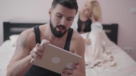 Retrato-de-hombre-caucásico-adulto-sentado-en-la-cama-y-jugando-videojuegos-usando-tableta.-Seductora-mujer-rubia-en-el-fondo-esperando-a-su-novio-o-marido.-Adicción-a-los-juegos,-problemas-matrimoniales.