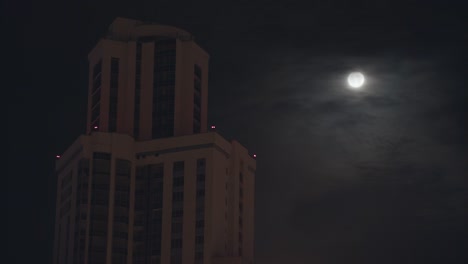 Night-cityscape-with-a-high-building-on-dark-sky-background.-Stock-footage.-Full-moon-on-dark-cloudy-sky