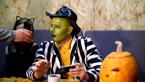 Halloween-party,-a-man-with-a-green-face-,-wearing-a-hat-and-a-girl-dressed-in-cat-costume-,-are-looking-into-the-phone,-reading-smth,-talking