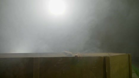 Young-teenager-female-zombie-waking-up-from-the-dead-getting-out-from-her-coffin-on-a-gray-smoke-background-with-fog-on-halloween-night