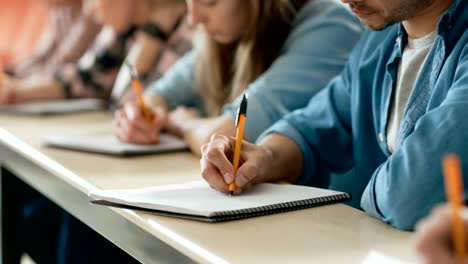 Filmfootage-aus-einer-Reihe-von-Multi-ethnischen-Schüler-im-Klassenzimmer-Prüfung-/-Test-Holding-Stifte-und-Schreiben-in-Notebooks-im-Fokus.-Helle-Jugendliche-Studie-an-der-Universität.