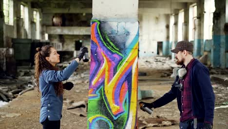 Creative-team-of-two-urban-painters-are-drawing-graffiti-with-spray-paint-while-decorating-old-industrial-warehouse-with-destroyed-dirty-walls-and-windows.