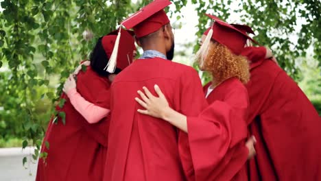 Fröhliche-Freunde-Absolventen-in-Kleidern-und-Mortarboards-sind-einander-nach-Abschlussfeier-gratulieren,-sie-umarmen-und-lachen.