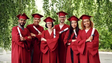 Portrait-of-group-of-graduating-students-holding-diplomas-and-standing-outdoors-with-arms-crossed-wearing-gowns-and-mortar-boards,-smiling-and-looking-at-camera.