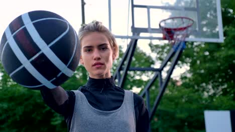 Schöne-blonde-Frau-zeigt-Basketball-in-Kamera,-Profi-Spieler-stehen-im-Park-tagsüber-Hoop-im-Hintergrund