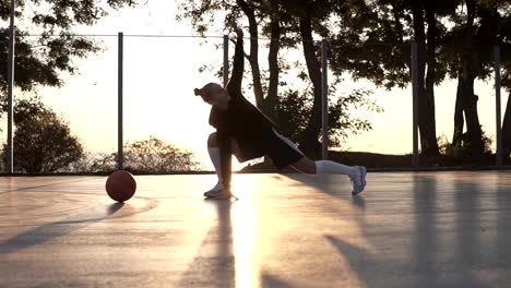 Joven-caucásica-forma-a-mujer-con-capucha-y-pantalones-cortos-calentando-en-la-mañana-en-cancha-de-baloncesto.-Estirar-las-piernas-antes-de-entrenamiento-de-baloncesto.-Al-atardecer-de-mañana