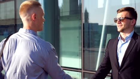 Two-young-businessmen-meeting-near-office-building-and-greeting-each-other.-Confident-colleagues-shaking-hands-and-discussing-work-in-urban-environment.-Concept-of-communication.-Close-up-Side-view