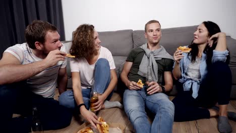 Essen,-Freizeit-und-Glück-Konzept---vier-lächelnde-junge-Menschen-zu-Hause-Pizza-essen-und-trinken-ein-Bier-auf-dem-Boden-sitzend.-Vorderansicht