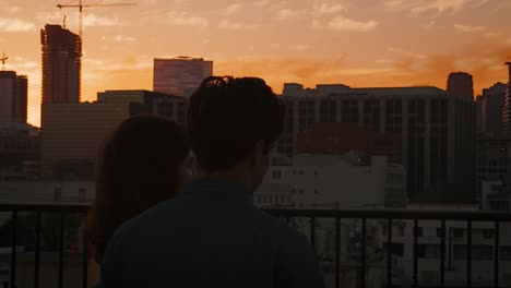 Rückansicht-des-Paares-auf-auf-dem-Dachterrasse-mit-Blick-über-Stadt-Skyline-bei-Sonnenuntergang