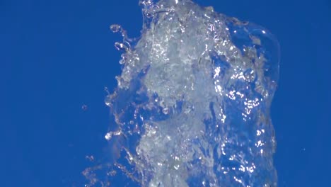 A-splash-of-clear-ice-water.-Closeup-of-light-splashing-water-in-sunny-fountain-in-park-in-summer-on-blue-sky-background.--Water-flow-of-fountain-fly-up-in-air-with-many-splashes.-Slow-motion.