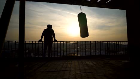 Boxer-on-terrace-after-training-looking-at-town,-bag-swinging,-beautiful-sunset,-sportsman-practicing,-power-training,-strong-guy-hard-exercising,-strength-exercises,-workout,-handheld,-sunny-day.