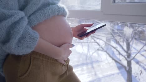 tecnología-móvil-moderno-para-las-mujeres-embarazadas,-maternidad-mujer-con-abdomen-grande-con-teléfono-inteligente-contra-la-ventana-en-la-luz-del-sol-en-día-de-invierno