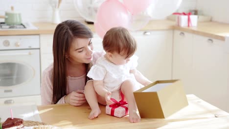 Neugierige-Mädchen,-die-auf-dem-Tisch-sitzt-und-die-Geschenkbox-an-ihrem-ersten-Geburtstag-öffnet,-ihre-liebevolle-Mutter-spricht-mit-ihr-in-der-Nähe