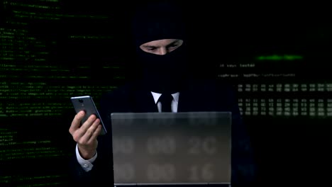 Criminal-in-suit-and-balaclava-breaking-security-code-using-laptop-and-phone