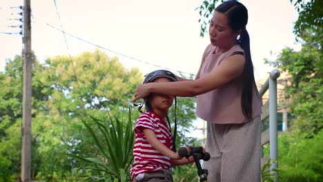 Madre-ayudando-a-su-hijo-(niño-de-pelo-largo)-a-ponerse-el-casco-de-la-bicicleta-al-aire-libre.