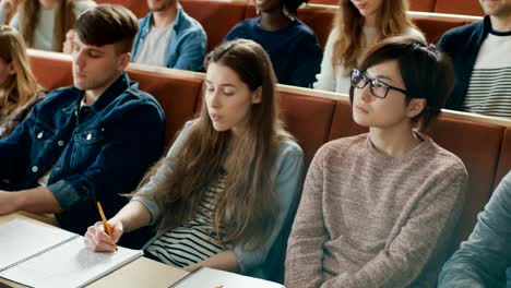 Kamera-mit-Blick-auf-Klasse:-College-Professor-hält-Vortrag-zu-einem-Klassenraum-voller-Multi-ethnischen-Studenten.-Dozent-Gesticulates-mit-seinen-Händen-während-Kursteilnehmer-hören.