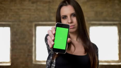 Young-pretty-attractive-women-with-long-hair-showing-phone's-display-into-camera,-green-screen-on-phone's-display,-brick-building-background