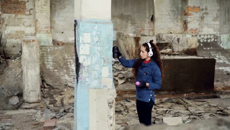 Serious-pretty-girl-is-busy-painting-graffiti-on-old-dirty-pillar-in-empty-industrial-building-using-spray-paint-while-listening-to-music-with-headphones.