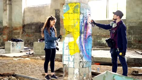 Slow-motion-of-skilled-graffiti-artists-bearded-guy-and-attractive-young-woman-working-together-in-abandoned-warehouse-decorating-damaged-column-with-abstract-image.