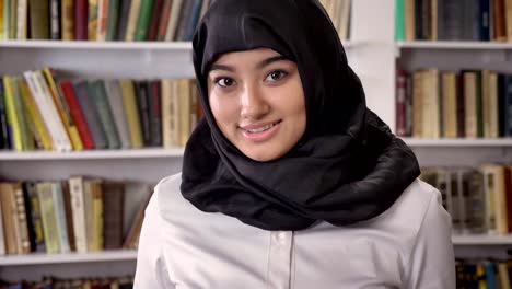 Joven-bonita-de-las-mujeres-musulmanas-en-hijab-mirando-a-cámara-y-sonriente,-de-pie-en-la-biblioteca,-estantes-con-fondo-de-libros