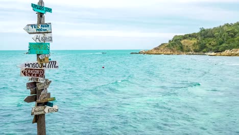 Wooden-direction-signpost-at-the-tropical-rocky-beach-with-ocean-waves-and-longtail-boat-in-background