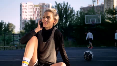 Jugador-de-baloncesto-femenino-feliz-hablando-por-teléfono-y-sonriente,-sentado-en-pista-al-aire-libre,-los-hombres-en-el-juego-de-fondo